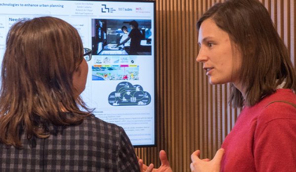 A woman in a red sweater explains a concept to another woman as they stand in front of a monitor displaying a poster