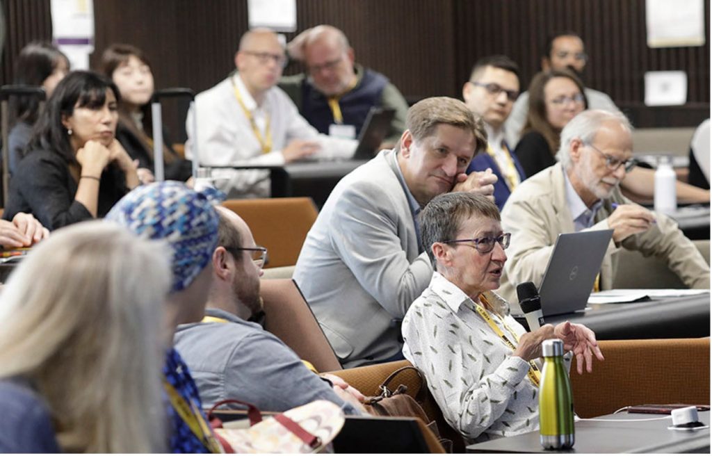 A group of people seated in a lecture hall, listening to Linda Newnes, who is seated in the front row. 