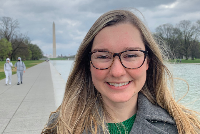 Morgan Orlovskiy, a woman with long blonde hair and glasses, smiles brightly at the camera. In the distance behind her is the Washington Monument, a tall obelisk.